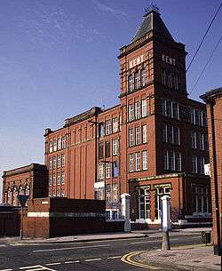 A mahogany-coloured four-storey brick factory is bathed in sunlight in front of a vivid azure sky. The view is from the ground looking up at a corner of the many-windowed cuboid building which is crowned by a tower upon which the text KENT appears in white lettering. In the foreground is a tarmac street leading the factory's entrance.