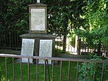 A picture of a grave, with trees, bushes, and a fence in the background and foreground. The grave itself has three markers, with carved text on it.