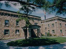 A flagpole and pine tree in front of a large two story stone building with many windows