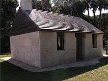 Color photo of the outside of a 2-room cabin covered in stucco, with a wood-shingle roof overtop and a chimney. One doorway in the middle of the longer wall indicates an entrance, and two glassless windows are located on both sides of the door.