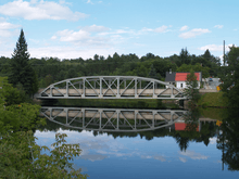 "A bridge crossing a river, the bridge reflects in the river. The steel girder bridge features an archway above the deck, with several beams interconnecting the arch with the deck.