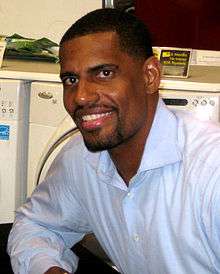 A black man wearing a blue button-down shirt sits and poses for a picture at a wooden table.