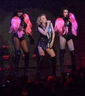 A woman in a sparkly vest is flanked by two background singers in black leotards with pink wigs as shoulder pads. All in the photo are singing into microphones.