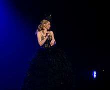 Kylie wears a feather dress with a gold corset on, holding a microphone looking towards her audience. She is also wearing a hat standing in front of a dark blue and black background (2011).