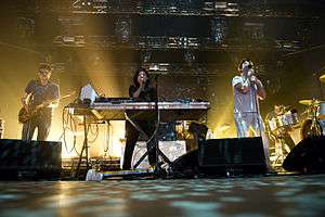 A man playing guitar, a woman playing the keyboard, and a man singing on stage