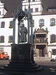 A statue of a man holding a book stands in front of a white building.