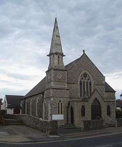 A pale stone building with a tiled roof sloping further on the left than on the right.  A tower with a two-light lancet window, diamond-shaped decoration and a short spire stands in the near corner.  A porch, with two doors and two windows in a lancet style, stands in front of the main body of the church, dominated by a four-light lancet.