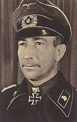 alt=A black-and-white photograph of a man wearing a black military uniform, peaked cap and a neck order in shape of an Iron Cross.