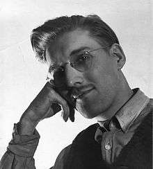 Black-and-white photograph of a man in glasses resting his head on his hand and smiling a the camera.