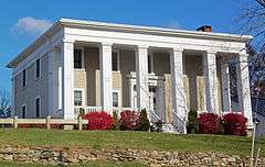 A two-story light brown wooden house with a flat roof and square columns in front on a rise in front of the camera