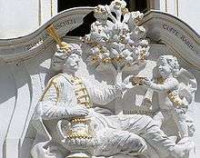 Relief of a young, cherub-like boy passing a cup to a reclining man with a moustache and hat. The sculpture is white with gold accents on the cup, clothes, and items.