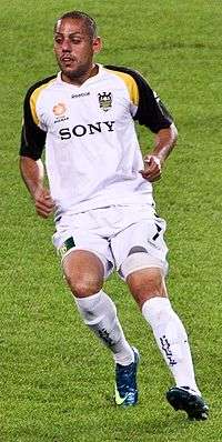 A man with very short hair who is wearing a white top, white shorts and wbite socks. He is standing on a grass field.