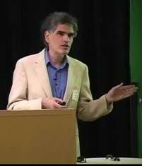 A man standing at a lectern in front of a blackboard, holding a slide clicker, and gesturing to the unseen audience