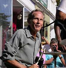 A man wearing a dress shirt, sitting behind a drum kit; spectators can be seen in the background.