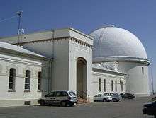 A white one story building with tall thin windows and an alcove entrance; at the right far end, a white domed building is present.