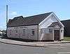 Three-quarter view of a long, low, cream-coloured painted hut with white cladding below the tiled roof.  An entrance porch with a brown door extends from the shorter side; the longer side, alongside a street corner, has four rectangular windows.  "The Lighthouse Community Church" is written in red letters above these.