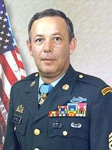 A colored photo of Littrell wearing his military dress uniform with ribbons and no hat. He is looking at the camera and an American flag is visible in the background.