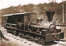 An old photograph showing a shiny black engine having a cab with open sides and a large, funnel-shaped smokestack