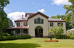 A pale yellow two-story house with reddish tiled roof in the Italian villa style. A pointed section projects from the front with the recessed, arched main entrance, and a tower pierces the roof in the rear.