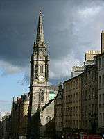 A line of gray 4 to 6 story row houses from the right, lead to a gray stone church with a single tower.