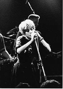 A black-and-white photograph of a woman singing into a microphone. Both her hands are raised and placed on the microphone stand. A man performing bass guitar is visible behind her, as well as drum cymbals and an overhead microphone; another woman's arm and the fretboard of an electric guitar are visible to her right.