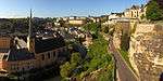 A panoramic view of a city. A small church is at the left, while a stony cliff flanks a small road leading to various buildings to the right.