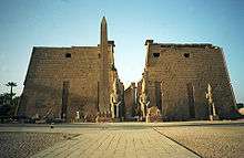 Pair of trapezoidal stone towers flanking a passage, beyond which a row of columns is visible. In front of the towers are several large statues and an obelisk.