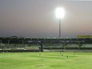 A cricketer bowling in front of largely empty stands. A single lit floodlight is visible in the background. Other cricketers can also be seen batting or fielding on the ground.