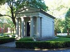 A brown stone building with a pointed roof and four columns in front in the middle of a paved circle and shrubbery. Across the top it says "Woodward".