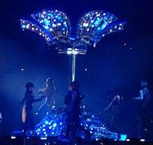 A bluish image of a pillar on a stage. It is flanked by women in horse-riding gear. From both the ends of the pillar hang a number of plates, the inside of which is lit by blue lights. One of the bottom plates has a number of stairs on which a similar horse-riding gear dressed lady, with her back to the image, is coming down.