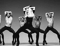 Black-and-white image of Madonna wearing tight black dress dancing in high heels. Behind her, a group of bare-bodied men emulate the moves, wearing similar high-heeled shoes