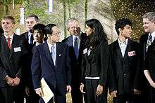 President George W. Bush and Japanese Prime Minister are on the right of a high school student. The high school student speaks. In the background are more high school students and important political figures.