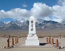 Photograph of a memorial at the Manzanar War Relocation Center. A low white obelisk with Japanese kenji characters and flower offerings stands against an expansive backdrop of the Owens Valley floor, snowy mountains, large clouds, and blue sky.