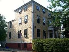 A three story yellow house with white trim, whose side faces the street. The porticoed entrance is behind a hedge.