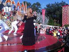 A woman wearing a long black gown. She has long golden hair and is holding a sparkling microphone. She is standing on a large red stage, surrounded by dancers in white attire. Additional background scenery include the audience and three background singers wearing white ensembles and standing on a large platform.