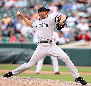 A baseball pitcher throws a pitch off a mound with his right arm. He wears a gray uniform and black baseball cap.