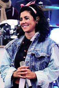 A young brunette woman, standing in front of a drumkit, holding a microphone and smiling. She is wearing a denim jacket, has a heart-shaped mark on her left cheek and has a pink bow in her hair.