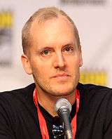 A man with thin brown hair, with a brown shirt, looking straight forward, with a microphone sitting in front of him.