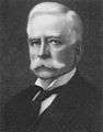 A headshot of a Caucasian male with a white handlebar moustache and white hair. He is wearing a white winged-collar shirt, black cravat and black coat.