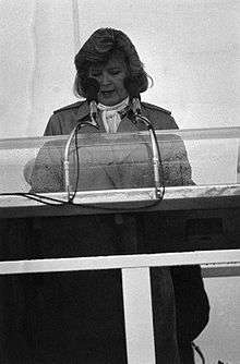 alt=A black and white photo of a woman in her fifties making a speech behind a podium and microphone