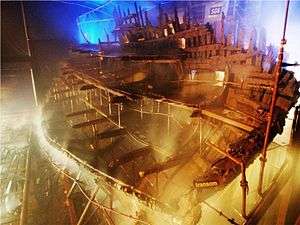 A diagonal section of a wooden ship seen from the stern inside a moodily lit building while it is being sprayed with water from a sprinkler system