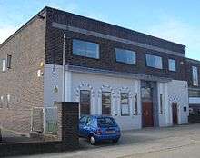 A brown-brick, flat-roofed, warehouse-style building. The lower half of its façade has been painted cream and decorated with arch-shaped window and door surrounds attached to the walls.  On the first floor there are three two-pane windows and one single window.  The side wall is mostly blank, with some small windows.