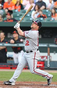 A stocky bearded man wearing a gray baseball uniform with red and navy-blue trim watches after following through on a swing