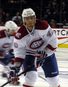 A Caucasian ice hockey player in his mid-twenties. He holds his hockey stick in a ready position while looking forward. He wears a white, visored helmet and a white jersey with red trim.