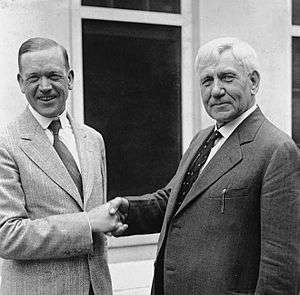 Two smiling middle-aged men in business suits shake hands in front of a window. They are standing but are visible only from the waist up.