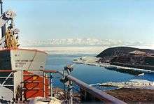  In left foreground are the upper works of a modern ship. To the right, on a low headland fringed with sea ice, isa square hut with a pitched roof. In the distance is a range of icy mountains