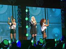 A young long-haired blonde woman singing into a microphone onstage. She wears a black skirt and black Bad Gal jacket. On her left and right are two brunette women dancing, each are wearing a sleeveless white top and leather shorts. A portraits of several bass speakers squared in pattern with background colors of neon green as the women's backdrop, as well as the iHeart Radio logo.