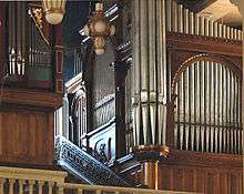 The metal pipes of two organs are in wooden cases of different dates and styles in the organ loft. To the side of the organ is a mosaic showing God as creator.