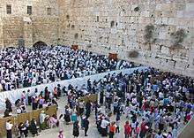 Two large groups of people, seen from slightly above them, separated by a white cloth barrier, standing before a beige stone wall whose top cannot be seen, with another wall in the rear. The group in the foreground is all female, the one in the rear is all male, with many wearing white robes or shrouds