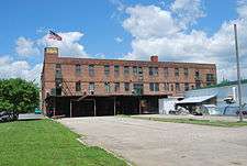 The same brick building seen from a different angle. An American flag flies from a short tower at the left end, and a flat roof projects over part of a parking area on that side at ground level. A one-story white building is at the right.